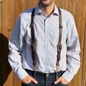 May include: A pair of dark brown leather suspenders featuring brass-toned metal buckles, D-rings, and swivel clips. The suspenders are worn over a white long-sleeved button-down shirt with a blue grid pattern and dark blue denim jeans.