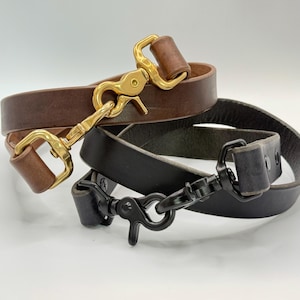 May include: Two leather straps, one dark brown and one black, each featuring a metal swivel snap hook. The brown strap has a polished gold-tone brass hook, while the black strap includes a matching matte black metal hook and two rivets.