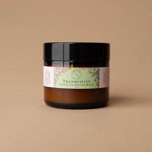 May include: A brown glass jar with a black lid. The jar has a label with the words "Tranquility Facial Clarifying Balm" and floral illustrations. The product is 1 oz.
