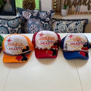 May include: Five personalized trucker hats with white fronts and mesh backs in green, orange, red, blue, and yellow. Each hat features a name, the text "King of Blackhead Hearts," and decorative embellishments.