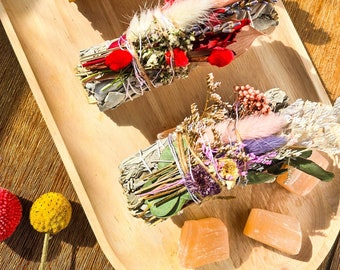 May include: Three bundles of dried herbs and flowers tied together with twine, arranged on a wooden tray with orange crystals. The bundles are made with sage, lavender, and other dried flowers.