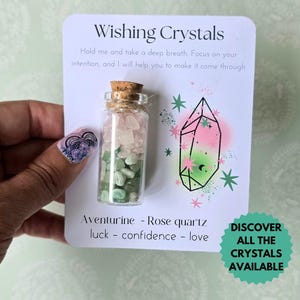 Peut inclure: Une petite bouteille en verre remplie de cristaux d'aventurine et de quartz rose, surmontée d'un bouchon en liège, est tenue dans une main. La bouteille est sur une carte avec le texte "Wishing Crystals" et un graphique de cristal.