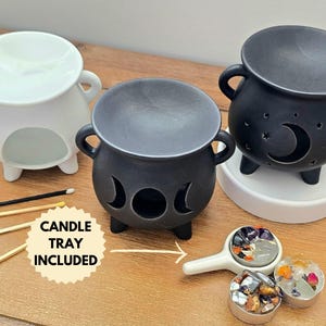 Puede incluir: Dos calderos de cerámica para calentar velas, uno blanco y dos negros, con un diseño de luna y estrellas. Los calderos están sobre una superficie de madera con una bandeja blanca y cerillas. "Bandeja para velas incluida" está escrito en una pegatina circular amarilla.