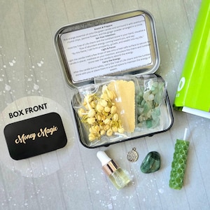 May include: A black metal box with the words "Money Magic" on the front. The box contains a green candle, a small bag of white flowers, a small bag of green crystals, a small bag of brown powder, a small bottle of oil, a silver charm, and a green stone.