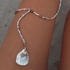 May include: A delicate, white beaded body chain with a pearlescent, teardrop-shaped pendant. The chain is composed of small, oval-shaped beads and larger, iridescent shell-like beads. The pendant is a large, iridescent shell piece.