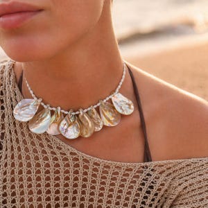 May include: A pearlescent shell necklace with a white beaded chain. The necklace features teardrop-shaped shells in shades of gold, white, and iridescent hues. The necklace is worn on a person's neck, with a beach background.