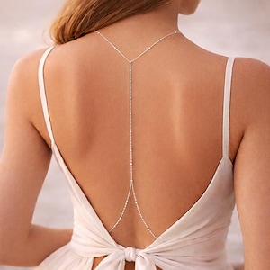 May include: A delicate silver body chain necklace. The necklace features a Y-shaped design that drapes down the back, with a series of small, sparkling accents. The necklace is worn over a white dress with spaghetti straps.