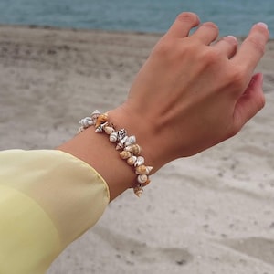 May include: A seashell bracelet composed of various small, natural seashells in shades of white, tan, and brown. The bracelet is worn on a wrist, with a yellow sleeve visible in the image. The background shows a beach and ocean.