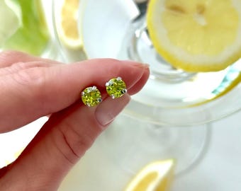May include: A pair of round, light green stud earrings with silver settings. The earrings are held in a hand, with a glass of water and lemon slices in the background. The earrings have a bright, gemstone appearance.
