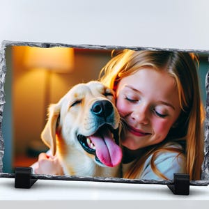 May include: A rectangular slate photo plaque with rough, chiseled edges, supported by two black plastic stands. The surface displays a colour photograph of a young person with long blonde hair smiling while embracing a light-coloured dog with its tongue out.