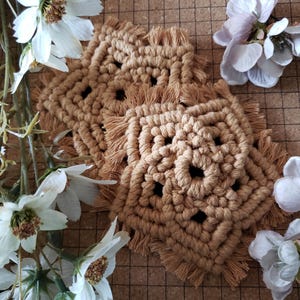 Puede incluir: Un juego de cuatro posavasos de macramé beige con un diseño geométrico y bordes con flecos. Los posavasos cuadrados parecen hechos a mano, con una estética natural y terrosa. Flores blancas decoran el fondo.