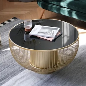 Könnte beinhalten: Ein goldfarbener Couchtisch aus Metall mit einer schwarzen Glasplatte. Der Tisch hat ein Gestell aus Drahtgeflecht und eine runde Form.