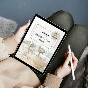 May include: A digital tablet displaying a graphic with the text "1000 Passive Income Ideas" and a starburst with the text "Make More Money". The tablet is being held by a person sitting on a couch.