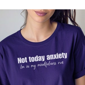 Puede incluir: Una camiseta de color púrpura oscuro con el texto "Not today anxiety" en negrita blanca y "I'm in my mindfulness era" en una fuente cursiva más pequeña. La camiseta tiene las mangas dobladas.