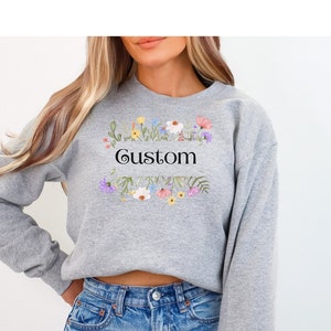 May include: A gray sweatshirt with the word "Custom" in black script, framed by a floral wreath. The sweatshirt features a small floral embroidery on the sleeve. The design incorporates colorful flowers and green leaves.