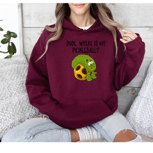 Könnte beinhalten: Burgunderfarbener Kapuzenpullover mit Fronttasche und Kordelzug. Der Kapuzenpullover zeigt den Text "DUDE, WHERE IS MY PICKLEBALL?" über einer Cartoon-Gurkenfigur, die einen gelb-braunen Pickleball umarmt.