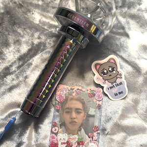 May include: A clear, diamond-shaped light stick with the word "SEVENTEEN" printed on the top. The handle has a rainbow prism design. A photo card and sticker are also present.