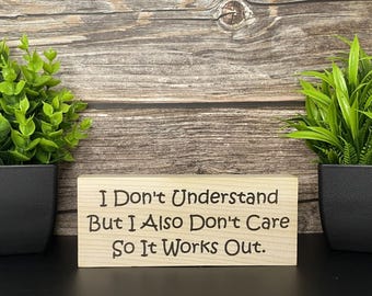 May include: A wooden sign with the engraved text "I Don't Understand But I Also Don't Care So It Works Out." The sign is light-colored and rectangular, with black text. Two potted green plants flank the sign.