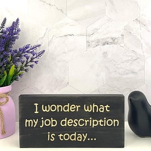 May include: A black wooden block with the text "I wonder what my job description is today..." in gold lettering. The block is on a white surface, with a pink vase of purple flowers to the left and a black decorative bird to the right.
