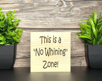 May include: A light-colored wooden sign with the text "This is a 'No Whining' Zone!" in a dark brown font. The sign is flanked by two potted green plants in black square pots.