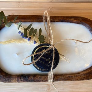 May include: A white candle in a wooden bowl with a small black and gold jar tied with twine and dried flowers. The jar has the text "Rustic Country Candles" on it.