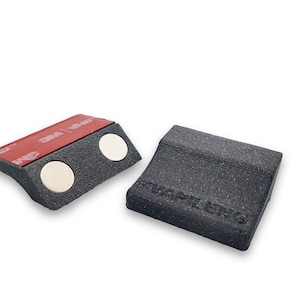 Può includere: Due supporti magnetici neri con retro adesivo. Un supporto ha due magneti bianchi e l'altro ha il testo "Fwapilens" stampato.