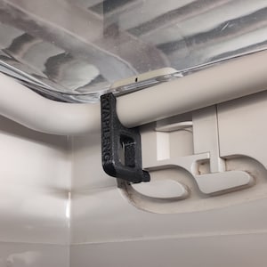 Puede incluir: Un pestillo de ventana de plástico negro con el texto "Knappleng" impreso en él. El pestillo está unido a un marco de ventana blanco.