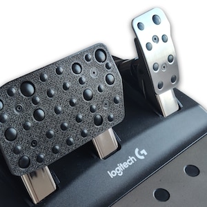 May include: A black and silver gaming pedal set. The foot pedals have textured surfaces with raised circular bumps. The silver pedals have circular indentations. The Logitech logo is visible on the base.