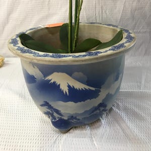 May include: A blue and white ceramic planter with a mountain scene and a decorative blue border. The planter holds an artificial orchid with white and yellow flowers and green leaves.