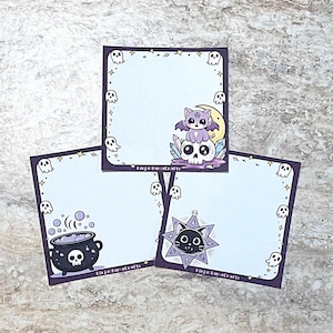 May include: Three purple and black square notepads with a white background and a different Halloween themed illustration on each. The first notepad has a purple cat sitting on a skull with a crescent moon behind it. The second notepad has a black cauldron with a skull and bones. The third notepad has a black cat inside a pentagram.