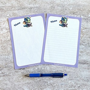 May include: Two purple-bordered notepads with the word "Notes" and a cartoon dragon illustration. One notepad is lined, the other is blank. A blue pen rests below the notepads. The notepads have the text "NinjaMamaCrafts" at the bottom.