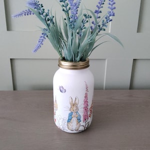 May include: A white mason jar vase with a gold lid, decorated with illustrations of Peter Rabbit and other characters. The vase holds faux lavender stems with purple flowers and green leaves. The vase is on a wooden surface.