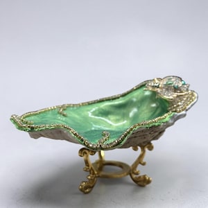 May include: A decorative, oval-shaped trinket dish with a light green interior and a gold-colored rim. The dish is supported by a gold-colored, ornate stand. The rim is encrusted with small, clear crystals and a few turquoise-colored stones.