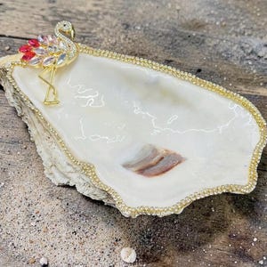 Puede incluir: Un joyero de concha de ostra con un borde de pedrería dorada. Un flamenco dorado con pedrería roja, rosa y transparente está en el lado izquierdo. El plato es blanco con un patrón marrón y beige.