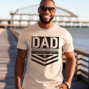 May include: A beige t-shirt with a black graphic that says "DAD DEDICATED AND DEVOTED To God, Family & Freedom" with a cross and chevron design.