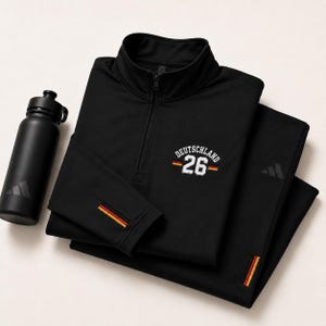 Könnte beinhalten: Schwarzes Sportbekleidungsset mit passender Wasserflasche. Das Set beinhaltet ein langärmliges Oberteil mit Reißverschluss und dem Wort "DEUTSCHLAND" auf der Brust sowie die Nummer "26". Ärmel und Hosenbein haben einen Streifen in den Farben der deutschen Flagge.