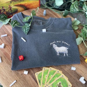 May include: A dark gray t-shirt with the text "Need any sheep?" embroidered on the front. A white sheep is embroidered below the text. The t-shirt is lying on a wooden surface with a red dice and several white wooden blocks.
