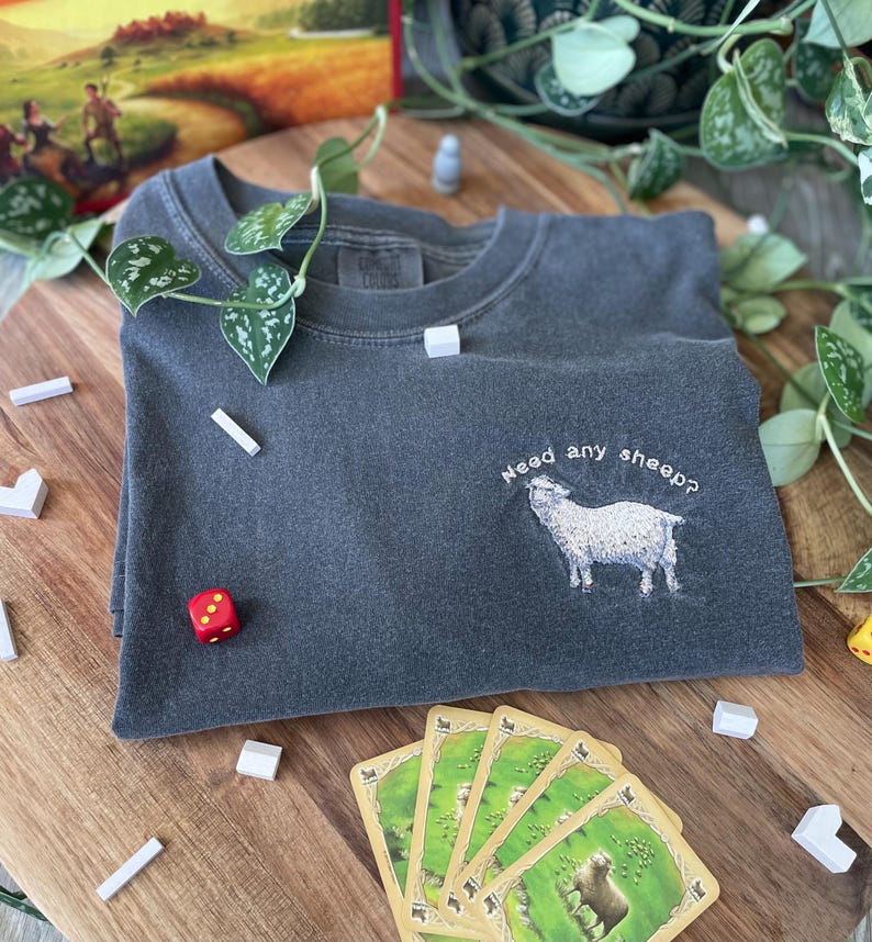May include: A dark gray t-shirt with the text "Need any sheep?" embroidered on the front. A white sheep is embroidered below the text. The t-shirt is lying on a wooden surface with a red dice and several white wooden blocks.