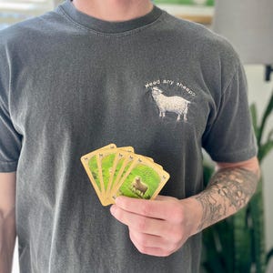 May include: A person wearing a dark gray t-shirt with the text "Need any sheep?" and a white sheep graphic. The person is holding a hand of five playing cards featuring a sheep graphic.
