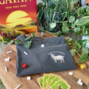 May include: A dark gray t-shirt with the text "Need any sheep?" embroidered on the front. The shirt is laying on a wooden surface with a game of Catan in the background.
