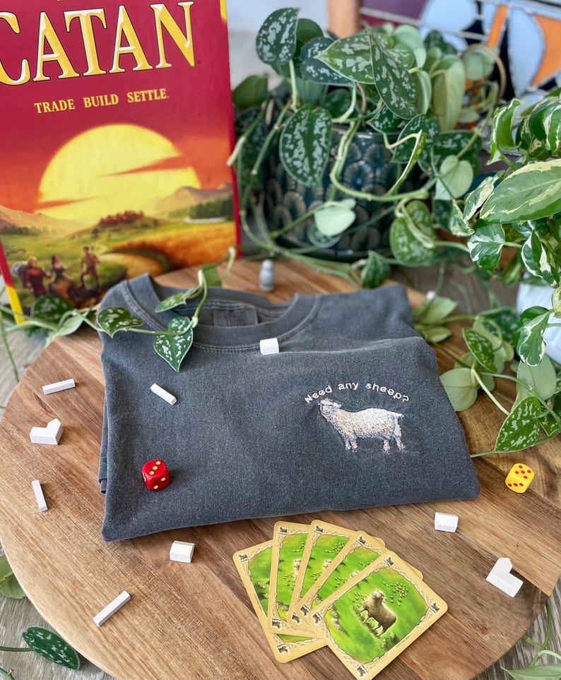 May include: A dark gray t-shirt with the text "Need any sheep?" embroidered on the front. The shirt is laying on a wooden surface with a game of Catan in the background.