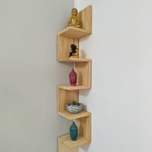 Könnte beinhalten: Ein Eckregal aus Holz mit fünf Ablagen. Die Ablagen sind versetzt und abgewinkelt. Die oberste Ablage hat eine goldene Buddha-Statue. Die zweite Ablage hat eine schwarze Buddha-Statue. Die dritte Ablage hat einen roten Buddha-Kopf. Die vierte Ablage hat eine kleine Topfpflanze. Die unterste Ablage hat einen türkisfarbenen Buddha-Kopf.