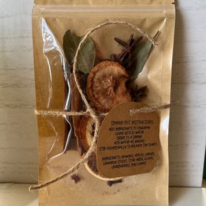 May include: A brown paper bag with a zipper closure contains a simmer pot mix. The mix includes oranges, apples, lemons, cinnamon sticks, star anise, cloves, cranberries, and bay leaves. The bag has a label with instructions for using the simmer pot mix.