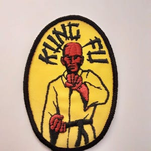 May include: Embroidered patch with a yellow oval background and a black border. The patch features a red-faced figure in a yellow uniform, black belt, and the words "KUNG FU" in black lettering.