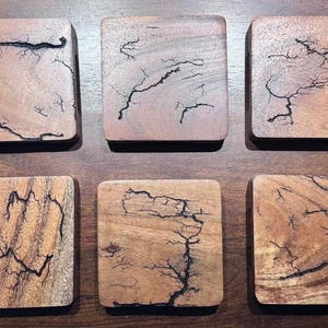 May include: A set of six square wooden coasters, each featuring a unique, dark, branching fractal pattern burned into the natural light brown wood grain. The coasters are arranged in two rows of three, displaying smooth surfaces and rounded corners.