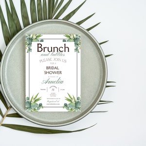 May include: A white invitation with green foliage border. The text reads "Brunch and bubbles, please join us for a bridal shower in honor of Amelia".
