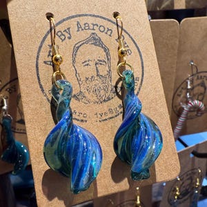 May include: A pair of blue and green glass earrings with a twisted design. The earrings are hanging from gold hooks and are labeled "By Aaron".