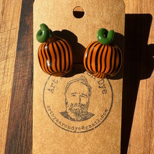 May include: A pair of pumpkin-shaped earrings. Each earring is orange with black vertical stripes and a green, swirled stem. The earrings are displayed on a brown tag with a circular stamp and the text "Art by Aaron Dye".