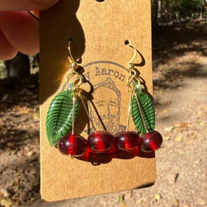 May include: A pair of dangle earrings featuring two dark red glass beads resembling cherries, paired with a green glass leaf bead with black stripes. The earrings are attached to gold-toned metal hooks and mounted on a brown cardboard display card.