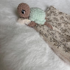 Puede incluir: Un peluche de tortuga hecho a mano con ganchillo, con cabeza y extremidades de color marrón claro, caparazón verde menta y una manta con estampado floral. La tortuga está sobre una superficie blanca y esponjosa.